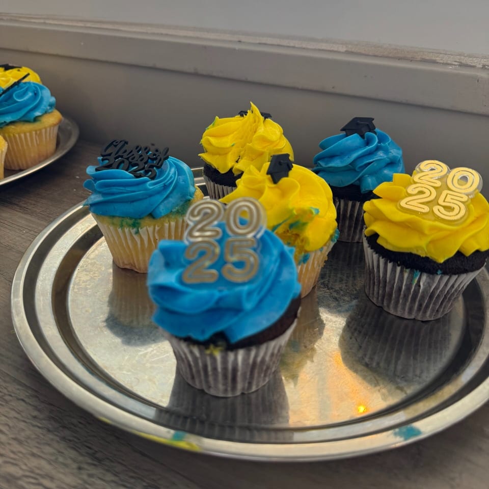 A silver tray holds blue and yellow frosted cupcakes, some with "2025" or graduation cap toppers.