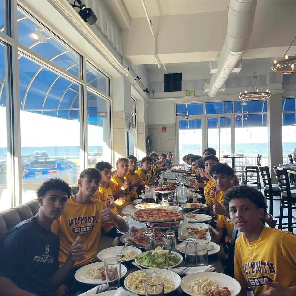 A group of young men in yellow shirts dining at a long table with pizza and pasta, ocean view.