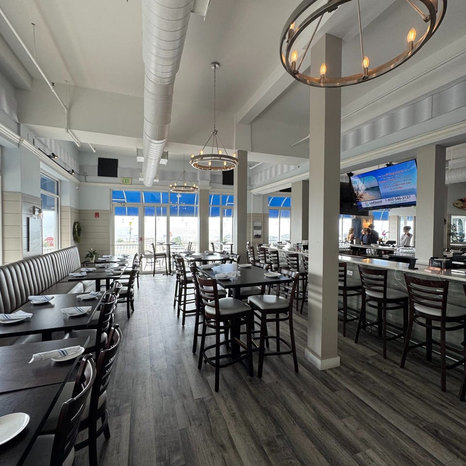 Spacious restaurant interior with dark tables, booth seating, a bar, and large windows overlooking water.