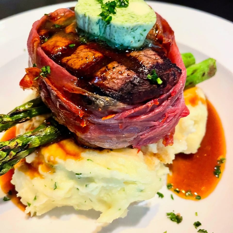 Prosciutto-wrapped steak with herb butter, mashed potatoes, asparagus, and gravy on a white plate.