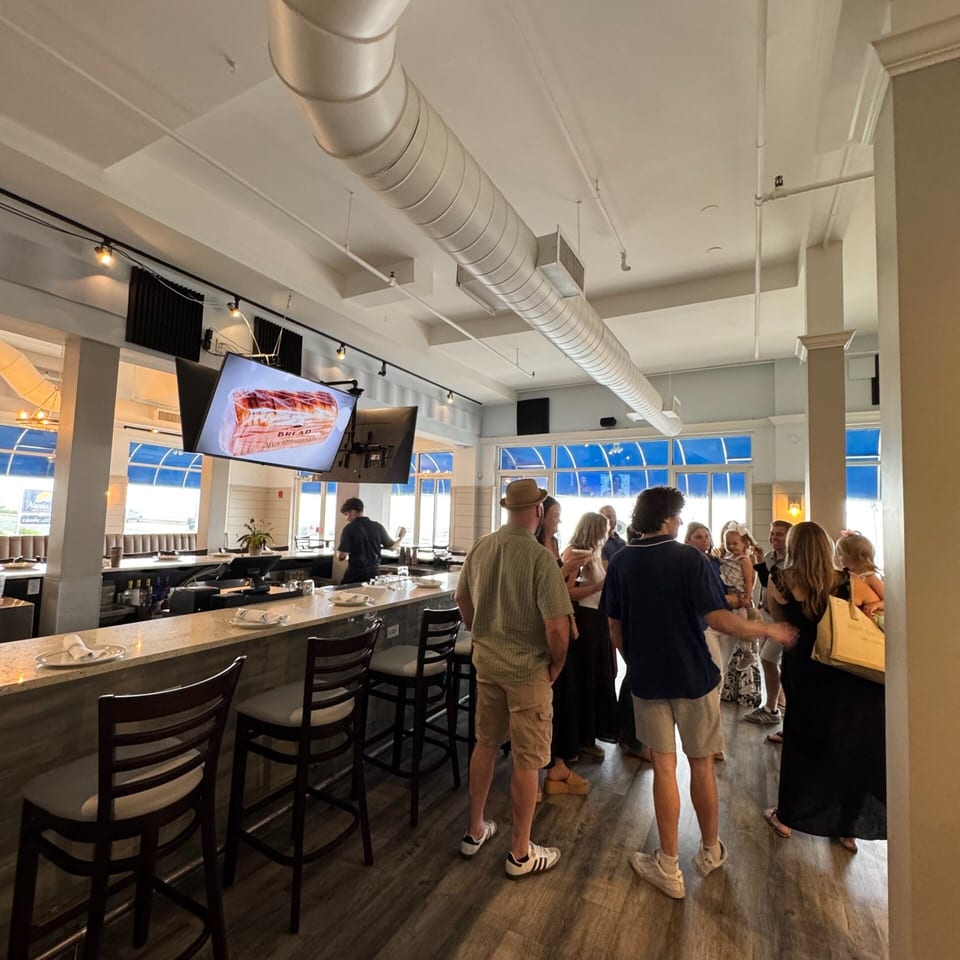 A lively restaurant interior with a long bar, bar stools, and patrons standing and conversing.