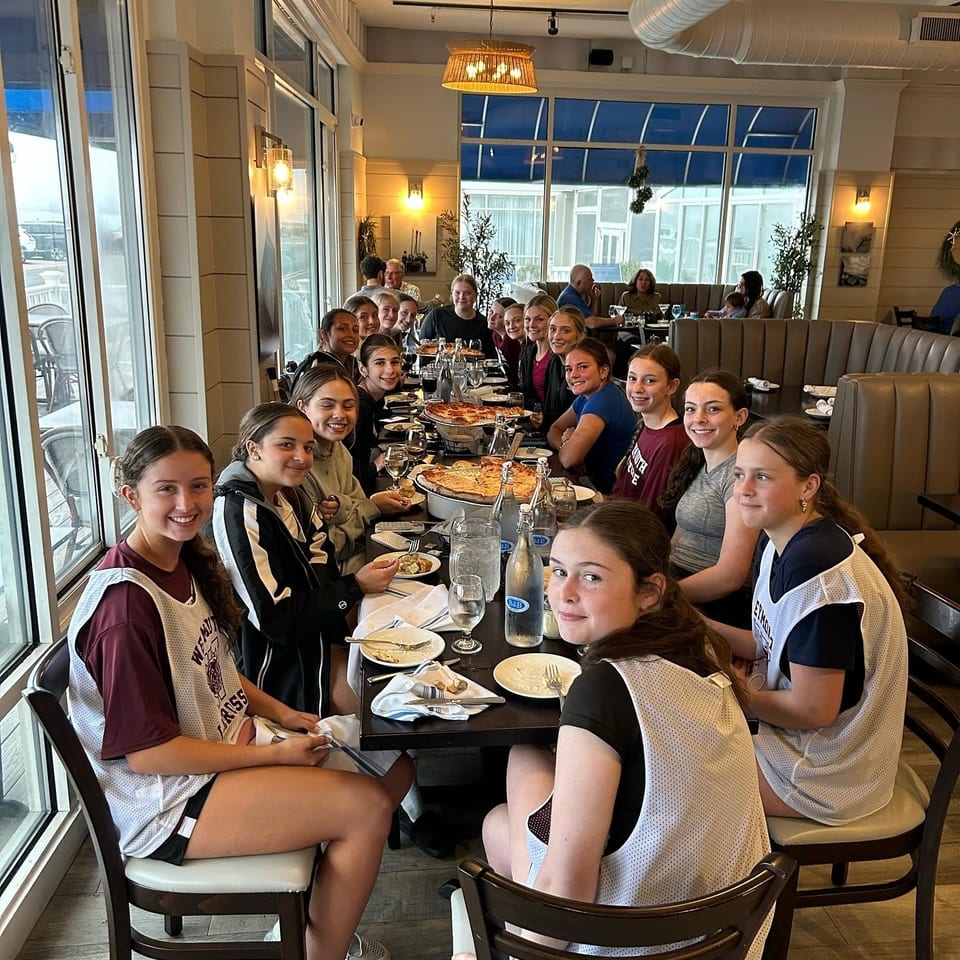 A large group of young women and girls smiling at a long table with pizzas in a bright restaurant.