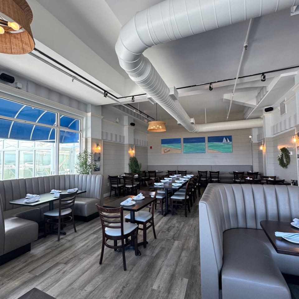 Spacious restaurant dining room with gray booths, dark tables, wood floors, and beach-themed art.
