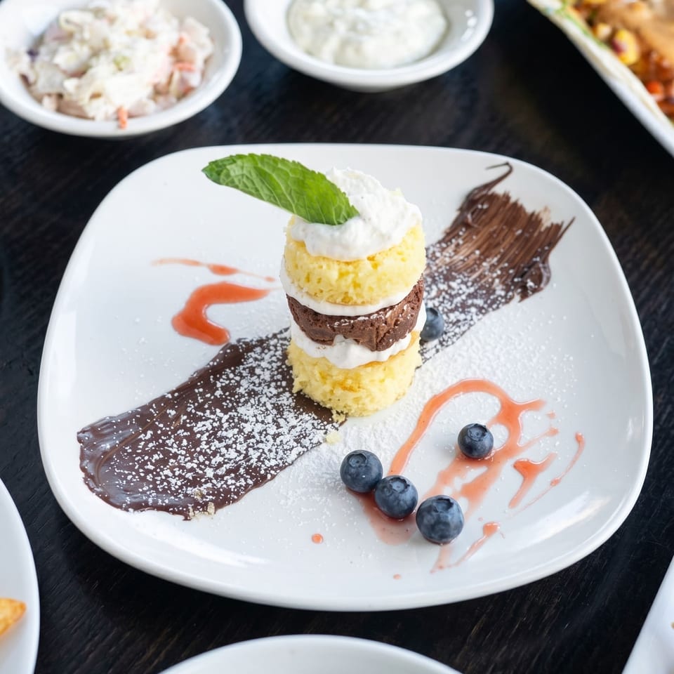 A layered dessert with yellow and chocolate cake, whipped cream, blueberries, and chocolate and berry drizzles.