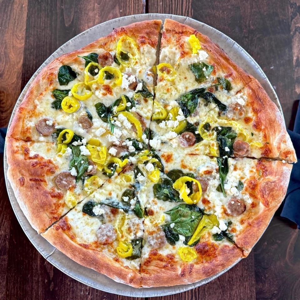 A whole pizza with white cheese, spinach, sausage, and yellow peppers on a dark wooden table.