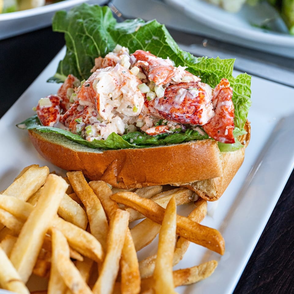 A lobster roll with lettuce and fries on a white rectangular plate, with other dishes blurred in the background.