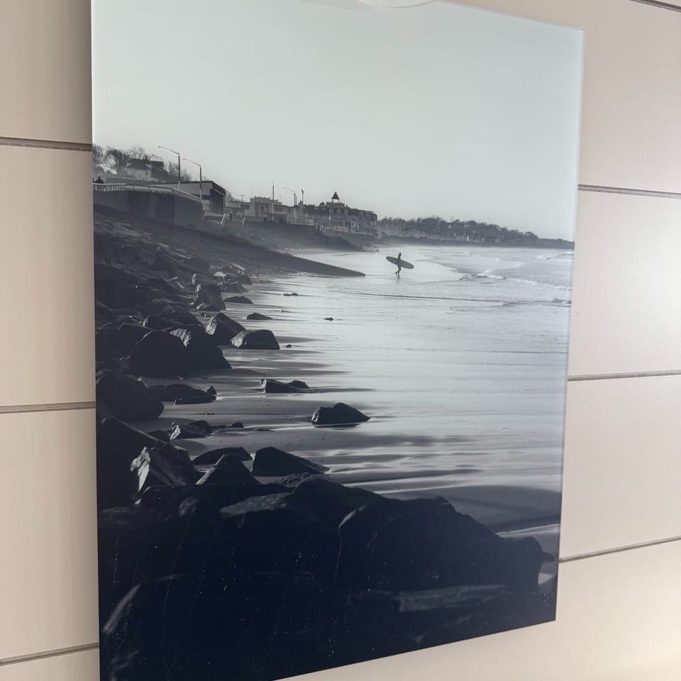 Black and white photo of a surfer carrying a board on a rocky beach with buildings in the background.
