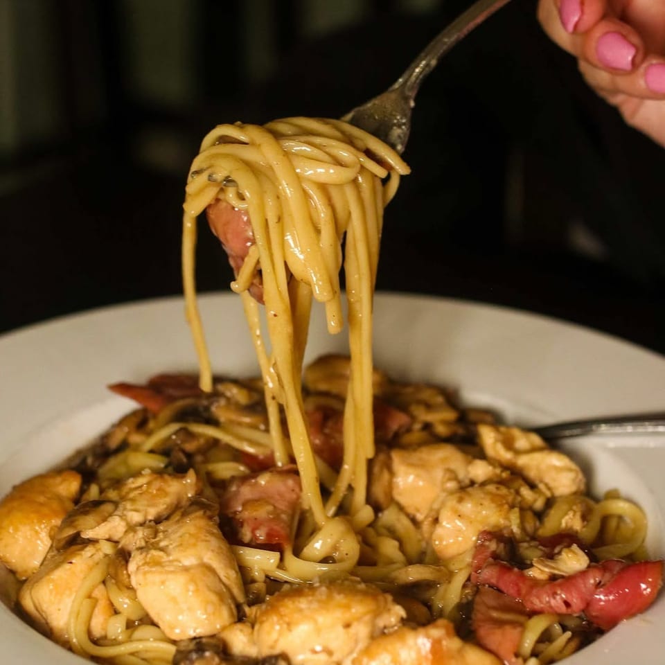 A fork lifts spaghetti noodles with chicken and bacon from a white bowl.