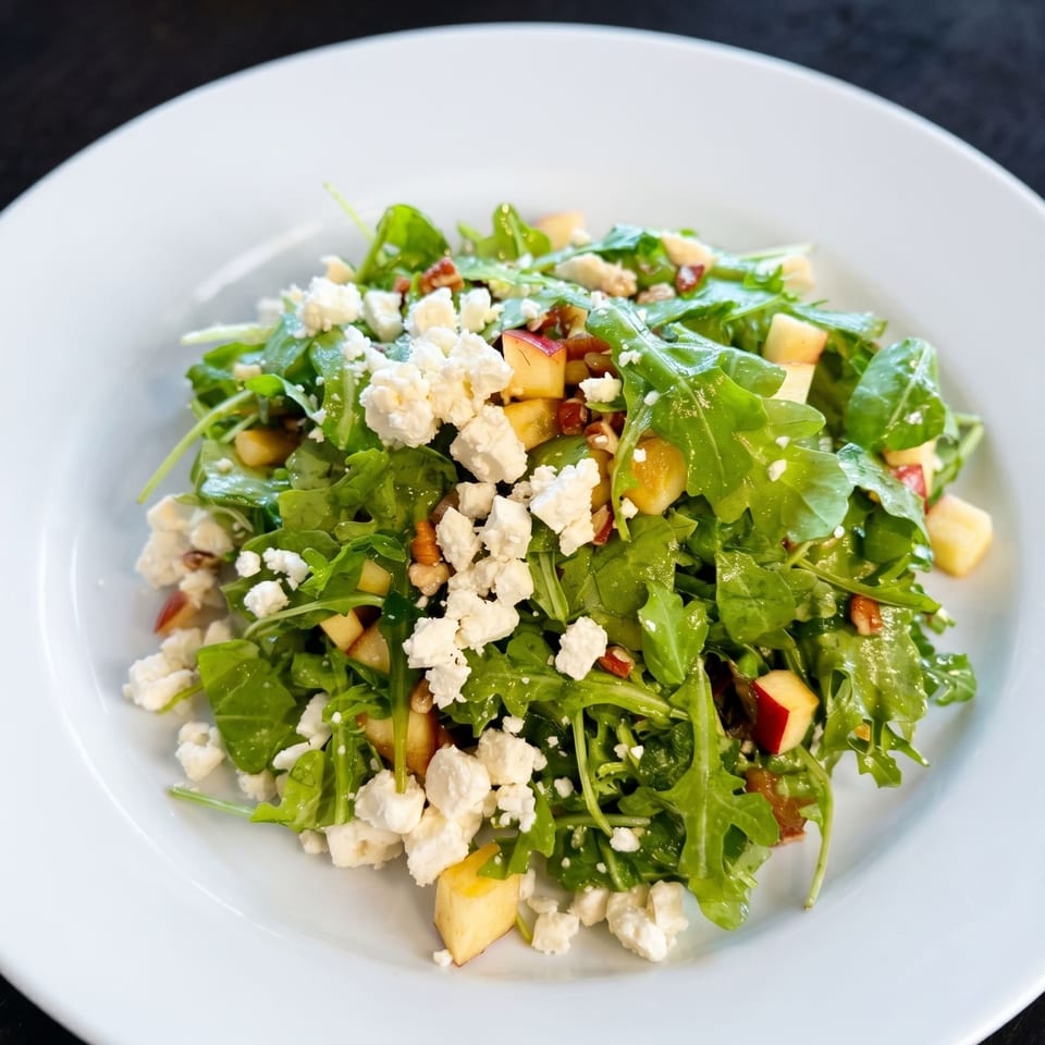 Arugula salad with crumbled feta, diced apples, and pecans on a white plate.
