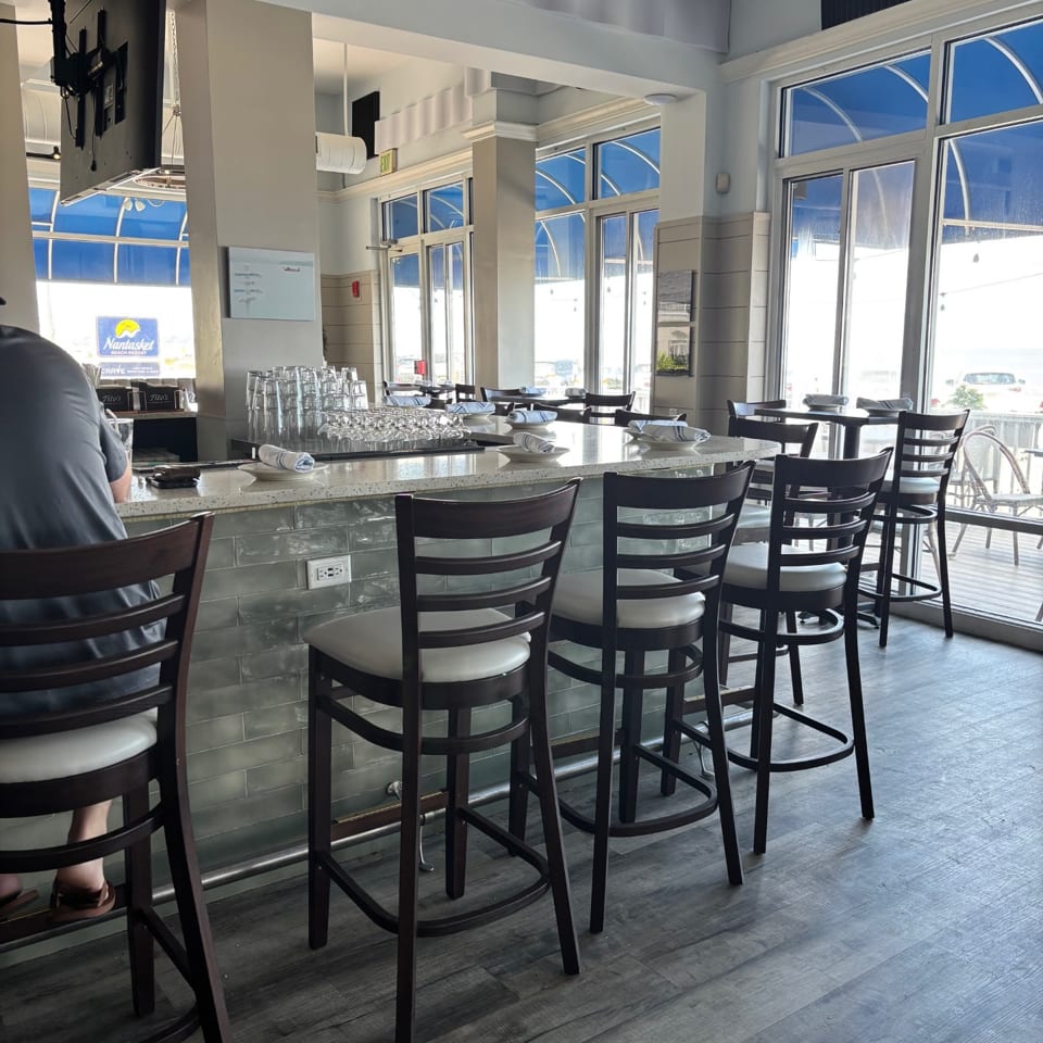 A bright, modern restaurant interior with a long bar, high stools, and large windows overlooking an outdoor patio.