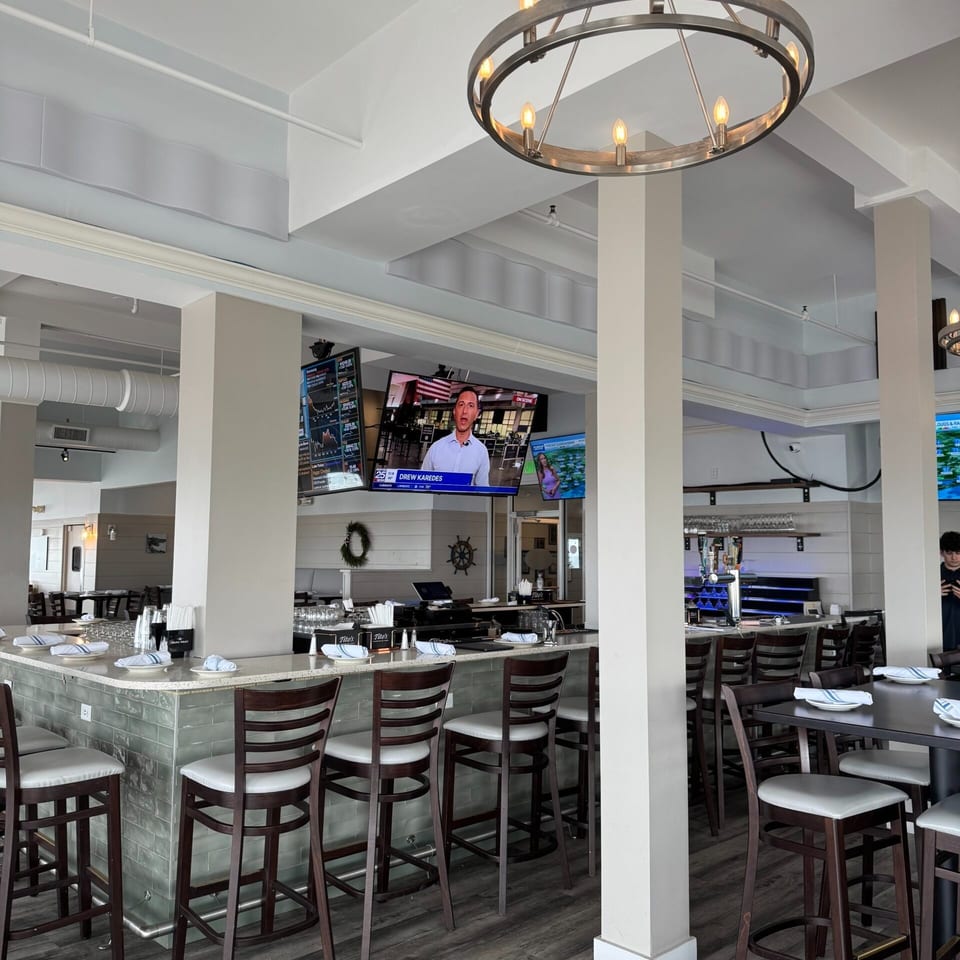 A modern restaurant interior with a long bar, high stools, and multiple TVs showing news and weather.